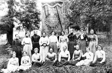 Bilde av gruppe kvinner utendørs. Bak dem en fane med teksten: "Det norske arbeiderpartis kvindeforening. Kvinder i alle land forener eder. Siftet marts 1895."