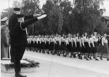 Kvinner  marsjerer i formasjon forbi uniformerte personer under en parade.