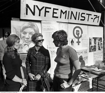 Gruppebilde 3 kvinner foran veggaviser og plakater, inkludert "Nyfeminist -71".