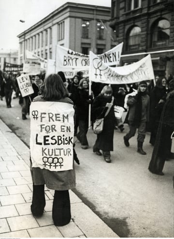 Demonstrasjonstog på Karl Johan i Oslo med paroler og plakater som "Lev lesbisk". I forgrunnen en kvinne med parole på ryggen: "Frem for en lesbisk kultur" dekorert med kvinnetegn.