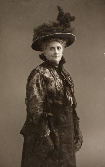 Svart-hvitt bilde av kvinne med kjole og hatt.