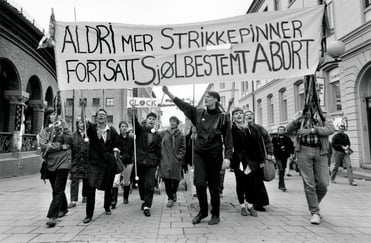 svart-hvitt bilde av en gruppe kvinner i demonstrasjonstog med et banner som lyder aldri mer strikkepinner fortsatt sjølbestemt abort.