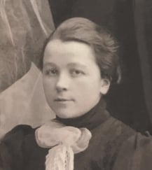 Historisk svart-hvitt portrettfotografi av en ung kvinne i mørke klær med en stor dekorativ sløyfe ved halsen.