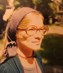 Fargeportrett av en kvinne med store briller og hodeplagg, fotografert utendørs.