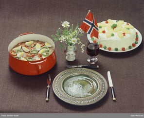 Bordoppdekning, tallerken, bestikk, gryte med gryterett med egg og paprikaringer, rødvinsglass, bløtkake, bolstervase med norsk flagg