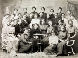 Gruppeportrett av rundt 25 kvinner i historiske klær, samlet rundt et bord i et studiofotografi fra tidlig 1900-tall.