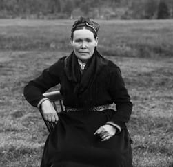 Kvinne i tradisjonell samisk drakt sitter på en stol utendørs i et historisk svart-hvitt fotografi.