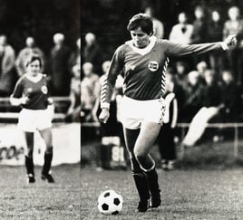 Fotografi av en fotballspiller i kamp, i ferd med å sparke ballen på banen. Nakne tilskuere og en annen spiller ses uskarpt i bakgrunnen.