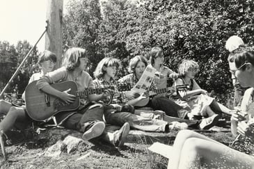 Gruppebilde av kvinner som sitter i graset og synger og spiller gitar. Sanghefte om Palestina.