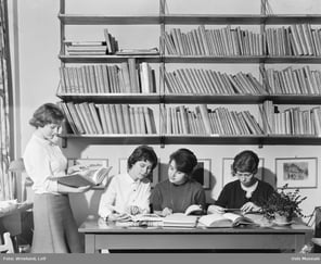 Fire kvinner leser og studerer bøker ved et bord foran store bokhyller i et bibliotek eller leserom.