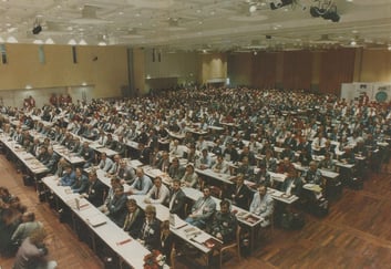 Stor konferanse eller kongress med hundrevis av deltakere samlet i en konferansesal med bord og møterekker.