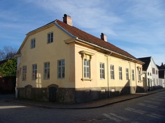 Gul trebygning i to etasjer ved et gatehjørne, med rødt tak og klassisk arkitektur.