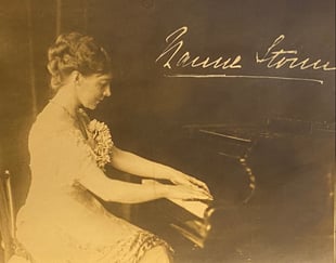Kvinne spiller piano i et historisk fotografi med signatur i bakgrunnen.