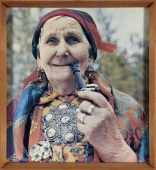 Eldre dame med fargerike klær, stor sølje og skaut ser mot kamera mens hun røyker pipe