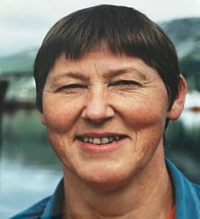 Portrettfoto av en kvinne med kort hår, fotografert utendørs med vann og landskap i uskarp bakgrunn.