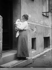 Kvinne holder et spedbarn utenfor inngangen til en bygård i et historisk svart-hvitt fotografi.