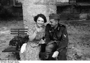 Person sitter på en benk sammen med en uniformert soldat utendørs. Soldaten holder en flaske, og begge er fotografert foran en steinbygning i et historisk svart-hvitt fotografi.