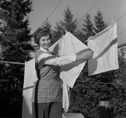 Historisk svart-hvitt-foto av en kvinne som henger opp senketøy på en klessnor utendørs, med trær i bakgrunnen.