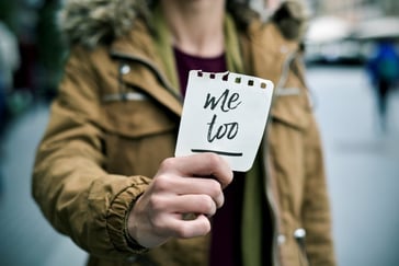 Person holder et kort med teksten «Me Too» fram mot kameraet, i et bymiljø med uskarp bakgrunn.