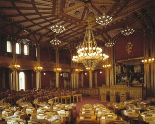 Bilde av en staselig sal med mørke trepaneler, høyt dekorert tak, flere lysekroner og rekker av pulter og stoler, som minner om et parlament eller bystyresal.