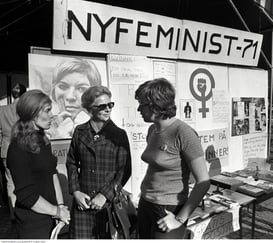 Gruppebilde 3 kvinner foran veggaviser og plakater, inkludert "Nyfeminist -71".