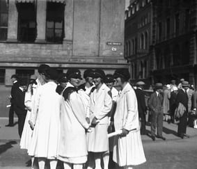 Historisk svart-hvitt-foto av en gruppe kvinner i lyse kapper og luer, samlet på et bytorg eller gatehjørne med bygninger og forbipasserende i bakgrunnen.