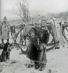 Historisk svart-hvitt-foto av en voksen med et barn i komse, stående i et snødekt landskap med slede, hund og gjerde i bakgrunnen.