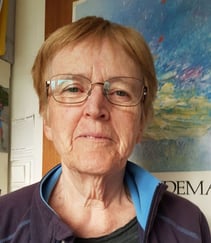 Et portrettfoto av en kvinne med briller, tatt innendørs foran en fargerik plakat eller et maleri.