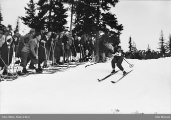 Et svart-hvitt bilde av en skiløper i fart under et skirenn, mens en gruppe tilskuere med ski står langs løypa i en skogkledd vinterkulisse.