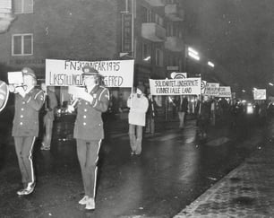 Historisk svart-hvitt-foto i en bygate, med korps-deltakere som bærer plakater med budskap om kvinners rettigheter, likestilling og fred.