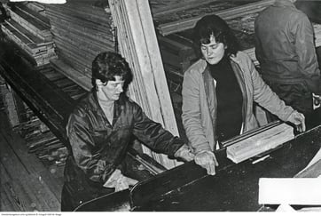 To kvinner arbeider med å skjære til plank i fabrikklokale. Mann i bakgrunnen.