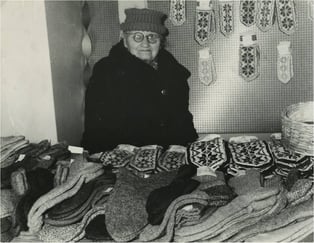Historisk svart-hvitt-foto av en kvinne som står bak et bord med strikkede votter og tekstiler utstilt for salg.