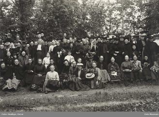 Historisk gruppefotografi av mange personer samlet utendørs i et skogsområde.