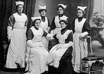Et gammelt svart-hvitt gruppeportrett av seks kvinner i sykepleieruniformer med forklær og hodeplagg, fotografert i et atelier.