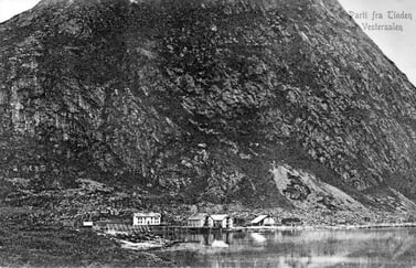 Historisk landskapsfoto av gårdsbygninger ved vannkanten, omgitt av et bratt fjell.