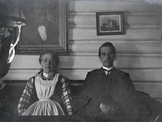 Historisk fotografi av et ektepar sittende innendørs foran en tømmervegg med innrammede bilder.