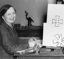 Historisk fotografi av en kvinne ved et arbeidsbord, omgitt av tegninger og kontorutstyr.