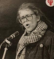 Kvinne med briller og skjerf taler i en mikrofon, med kvinnebevegelsens symbol synlig øverst i bildet.