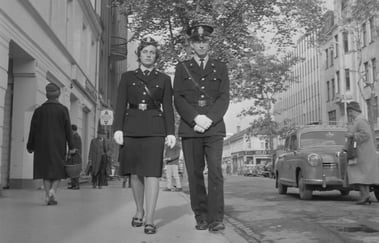 To uniformerte personer, en kvinne og en mann, går side om side i en bygate i et historisk svart-hvitt fotografi.