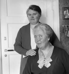 To kvinner poserer sammen innendørs i et svart-hvitt fotografi.