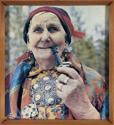 Eldre dame med fargerike klær, stor sølje og skaut ser mot kamera mens hun røyker pipe
