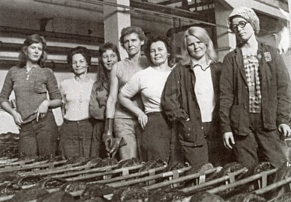 Et svart-hvitt gruppebilde av flere kvinner som står i et industrielt miljø, med metallgjenstander i forgrunnen.
