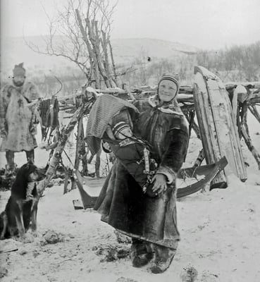 Historisk svart-hvitt-foto av en voksen med et barn i komse, stående i et snødekt landskap med slede, hund og gjerde i bakgrunnen.