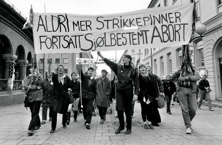 svart-hvitt bilde av en gruppe kvinner i demonstrasjonstog med et banner som lyder aldri mer strikkepinner fortsatt sjølbestemt abort.