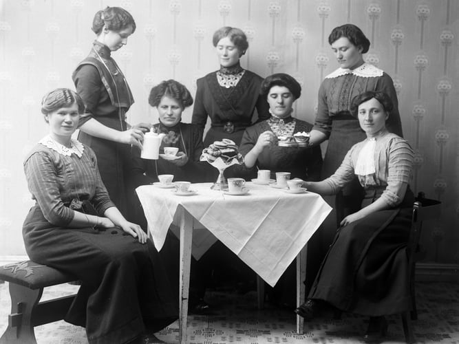 Seks kvinner samlet rundt et bord med kaffekopper og kaker i et historisk svart-hvitt fotografi.