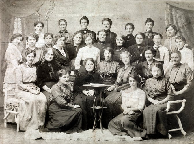 Gruppeportrett av rundt 25 kvinner i historiske klær, samlet rundt et bord i et studiofotografi fra tidlig 1900-tall.