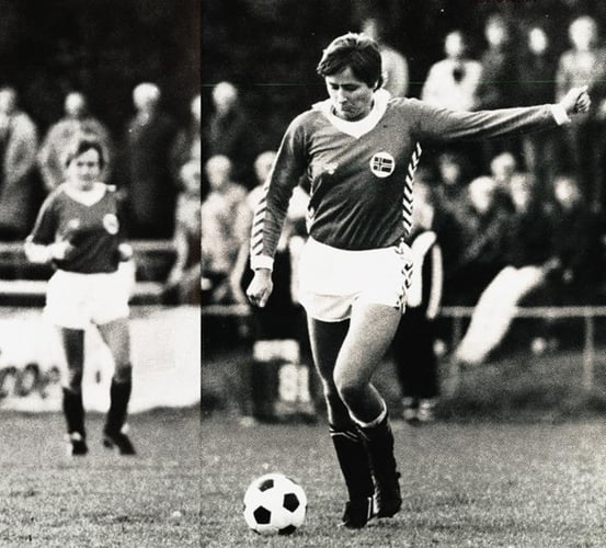 Fotografi av en fotballspiller i kamp, i ferd med å sparke ballen på banen. Nakne tilskuere og en annen spiller ses uskarpt i bakgrunnen.