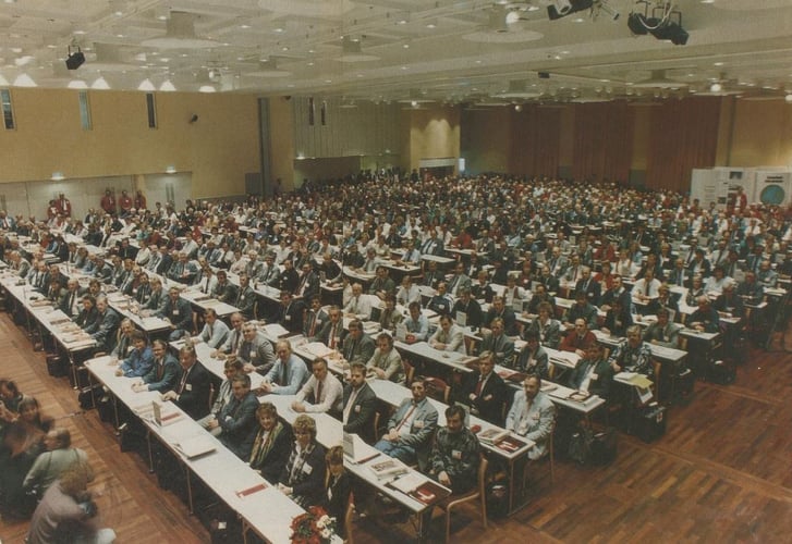 Stor konferanse eller kongress med hundrevis av deltakere samlet i en konferansesal med bord og møterekker.