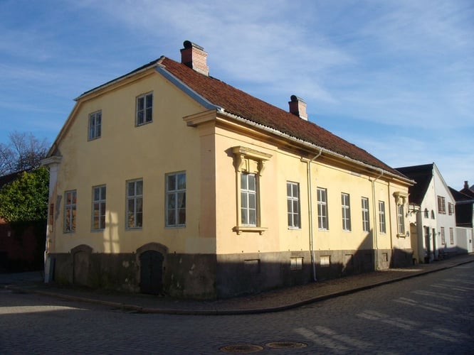 Gul trebygning i to etasjer ved et gatehjørne, med rødt tak og klassisk arkitektur.
