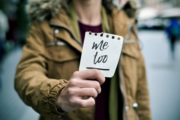 Person holder et kort med teksten «Me Too» fram mot kameraet, i et bymiljø med uskarp bakgrunn.